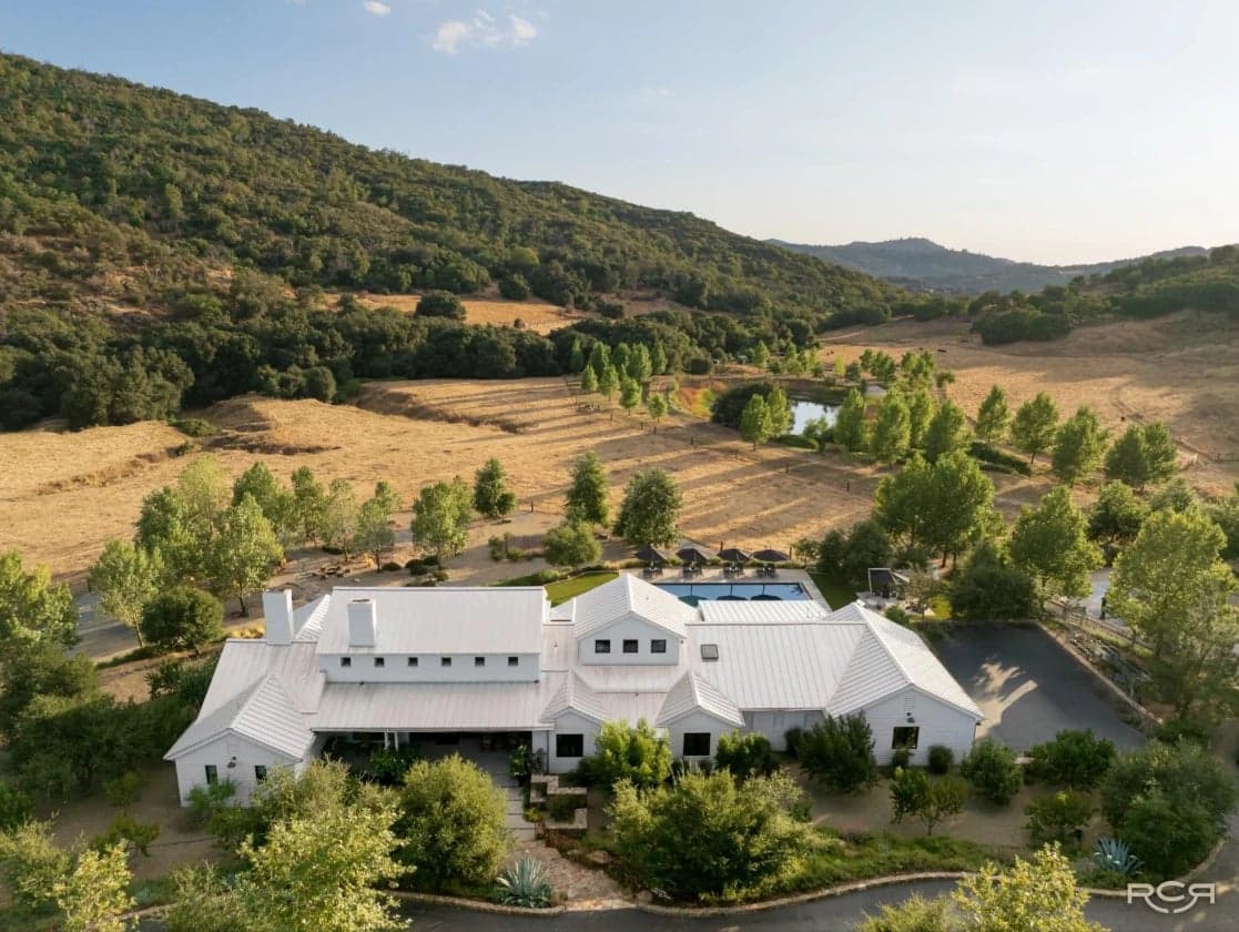 Aerial view of Red Cloud Ranch in Santa Ysabel, San Diego County: white ranch house with metal roof, pool, pond, and golden hills with oak-covered slopes